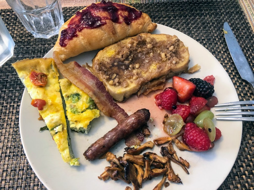 Close up of a plate covered with food: french toast with nuts, croissant with jam, two triangular slices of egg frittata, a slice of bacon, a link of sausage, a pile of colorful fruit, and a pile of orange mushrooms.