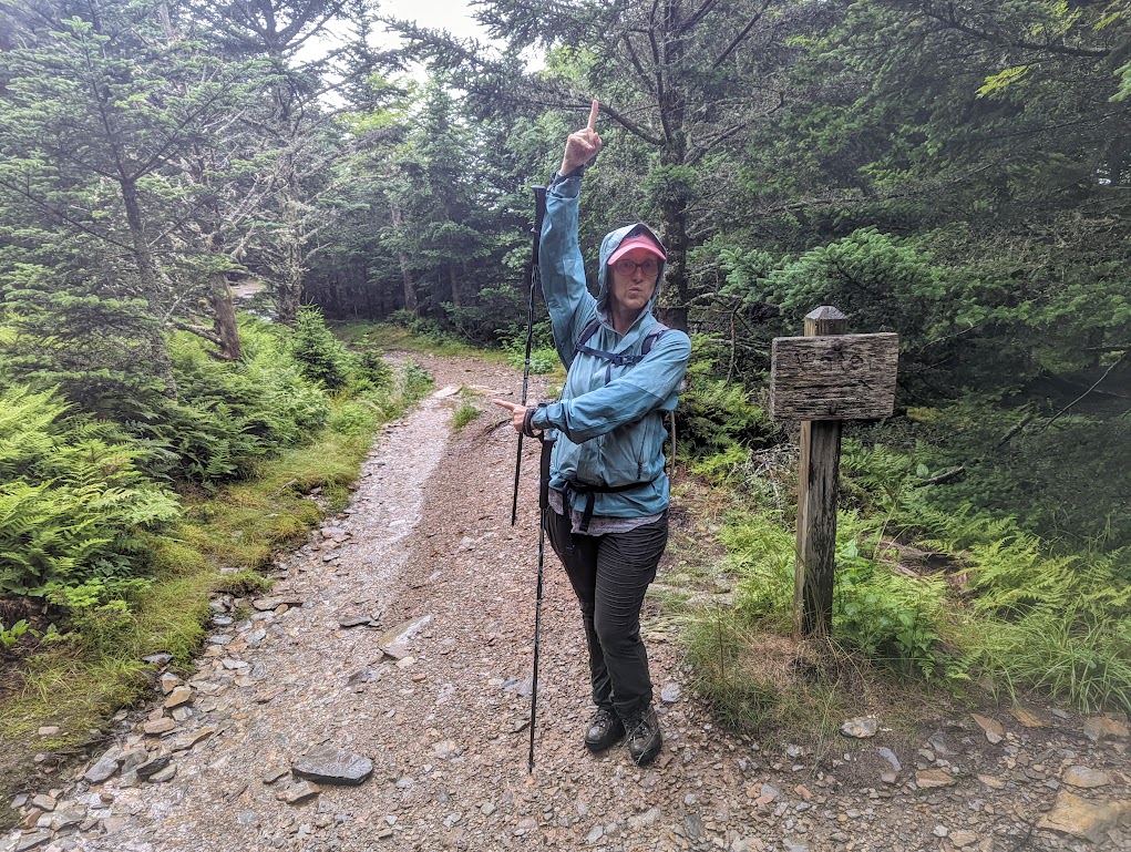 Laura stands beside a sign that says "WATER  [right arrow]" Laura is pointing in two other directions at all the rain coming down.