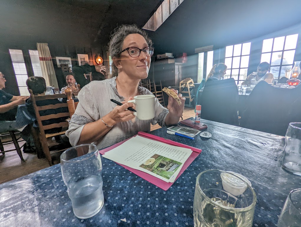 Laura holds a mug of hot chocolate. Journal pages are on the table in front of her and a crowded dining room is the background.