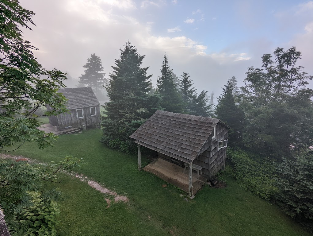 Two plank cabins perch on a grassy slope with trees, surrounded by fog. The sky directly overhead is blue.