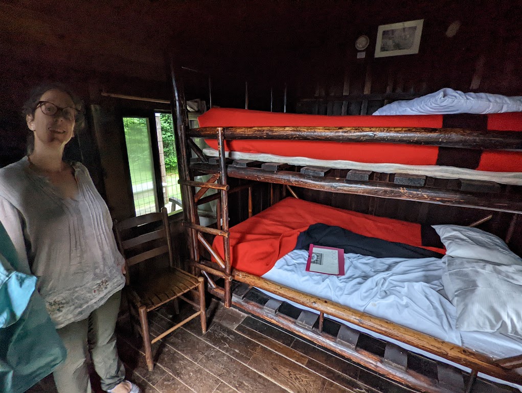 Laura, left, stands next two a bunk bed made up in red blankets inside a small, dark cabin.