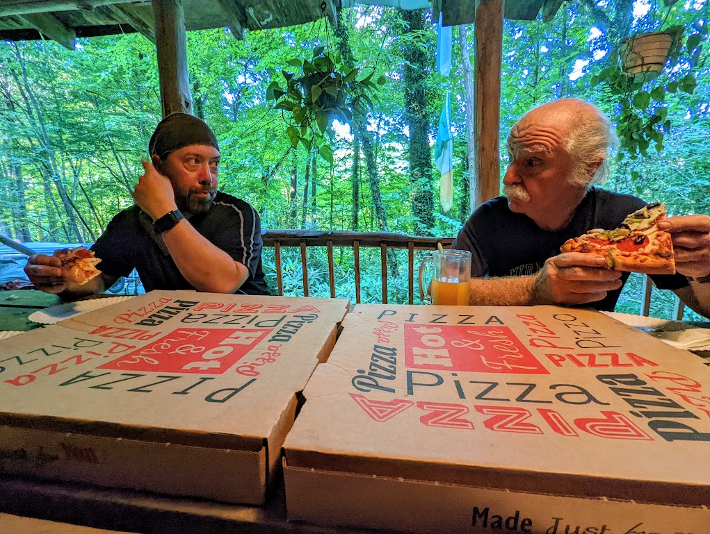 Two closed pizza boxes fill the entire bottom half of the frame. Behind the boxes (which is actually across the table), Hobz sits to the left and Bob Peoples to the right. Each holds a slice of pizza. They are looking at each other, both wearing incredulous expressions. Green trees can be seen behind them.