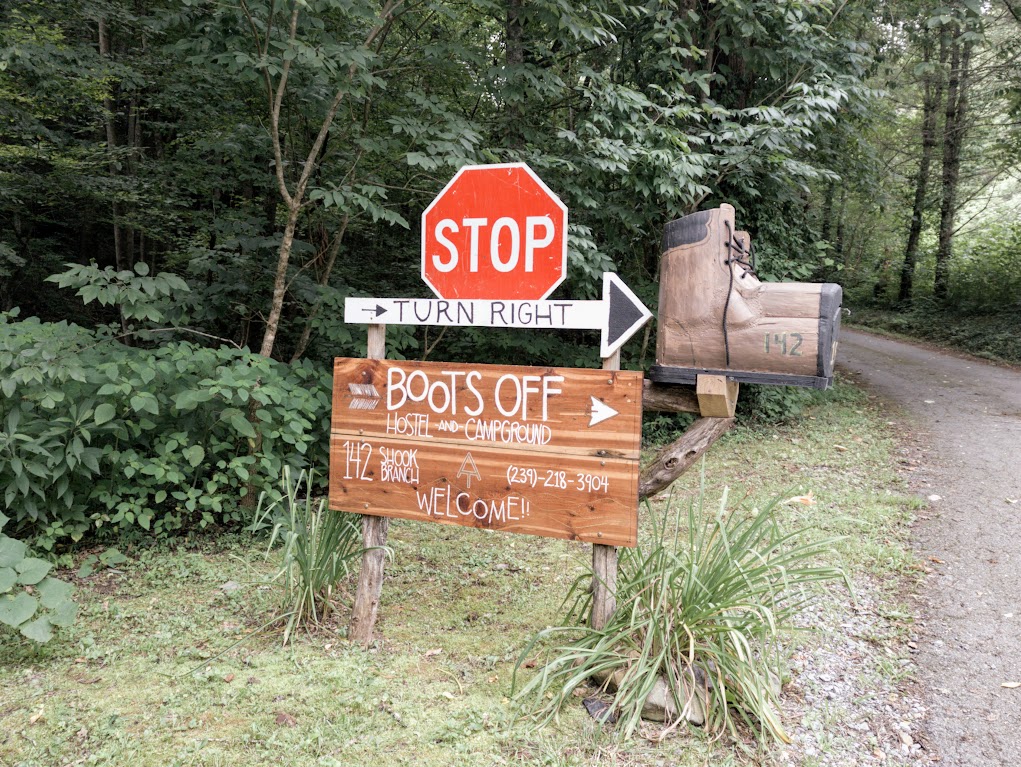 A roadside sign. At the top is a standard STOP sigh. Below it is a white arrow saying "TURN RIGHT". Below that is a sign that says, 

BOOTS OFF 
Hostel & Campground
142 Shook Branch
(239)-218-3904
WELCOME!!