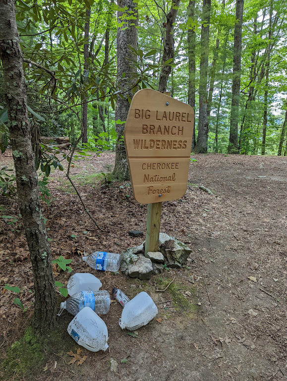 A vertical photo of the forest, with a sign posted on a wooden pole in the middle. It reads:
BIG LAUREL BRANCH WILDERNESS
CHEROKEE National Forest
Six large plastic bottles are clustered around the bottom of the sign.