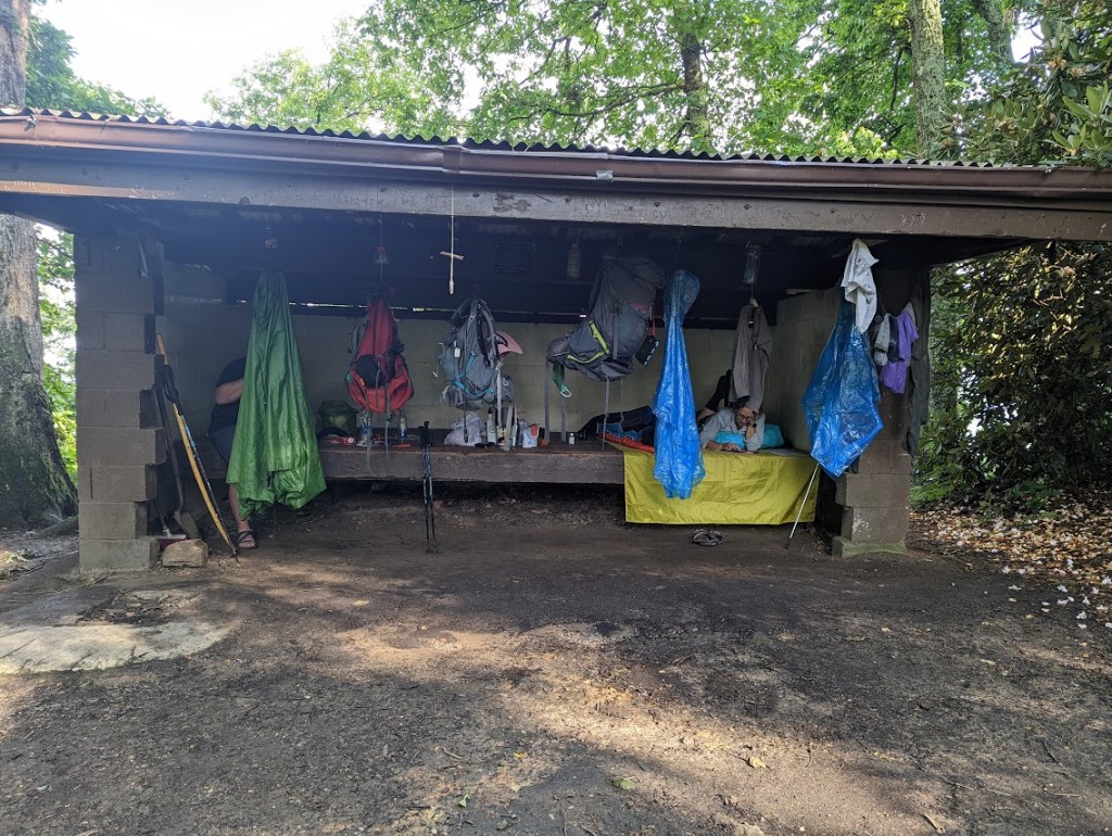 A photo looking into the shelter with gear, clothing, and backpacks hanging from every hook. Hobz and Laura can almost be seen behind all of it.