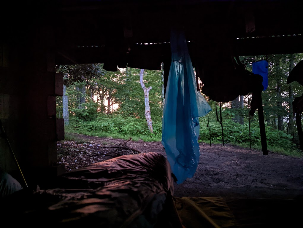 A photo taken from inside the shelter. The sleeping bag the photographer is in can be seen a the bottom. Plastic rain ponchos hang from the ceiling, with trees and a pink sky in the background.