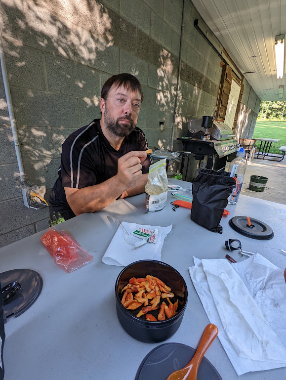 Hobz sits at a picnic table with a pot of chicken-of-the-woods mushrooms in front of him. He is holding up one piece of mushroom, looking very serious. 