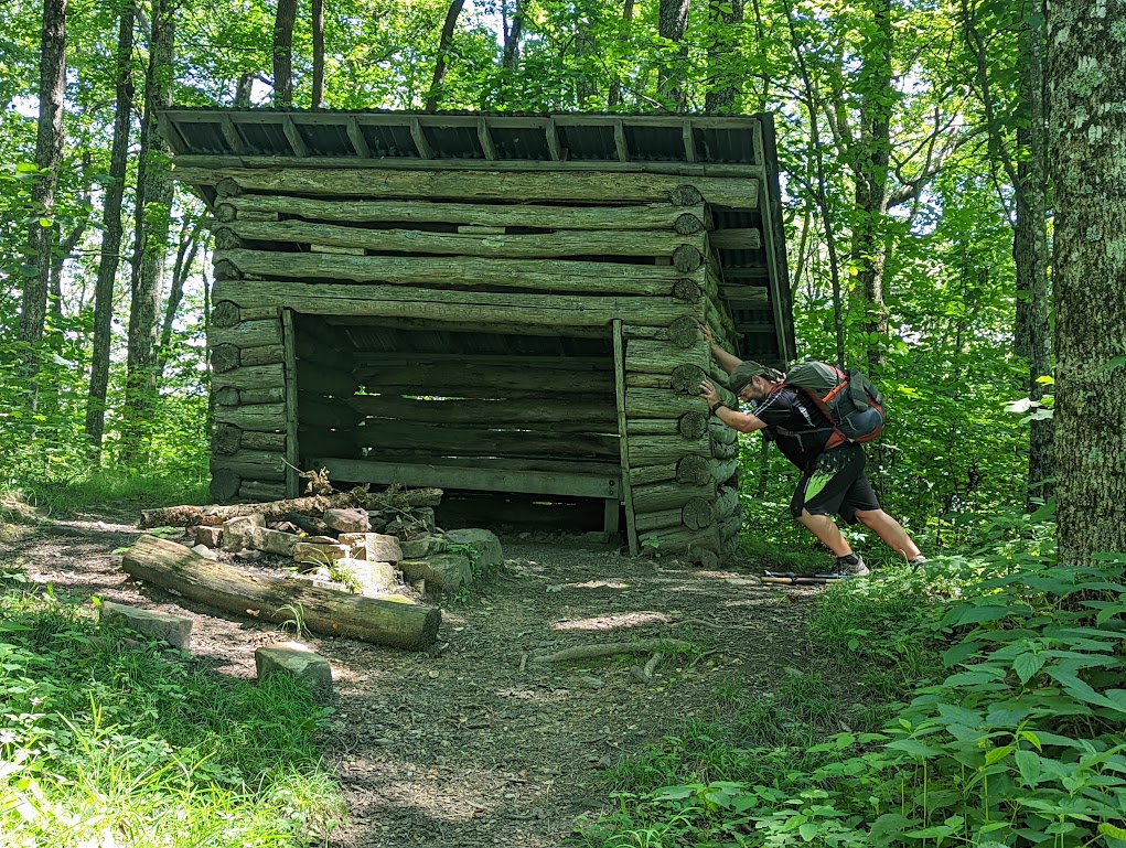 A log lean-to fills the frame. There are large gaps between each log and it appears to be listing to the left. Hobz pushes on the right side of the shelter, his feet braced as if making a huge effort, appearing to be responsible for the shelter's tilt. 
