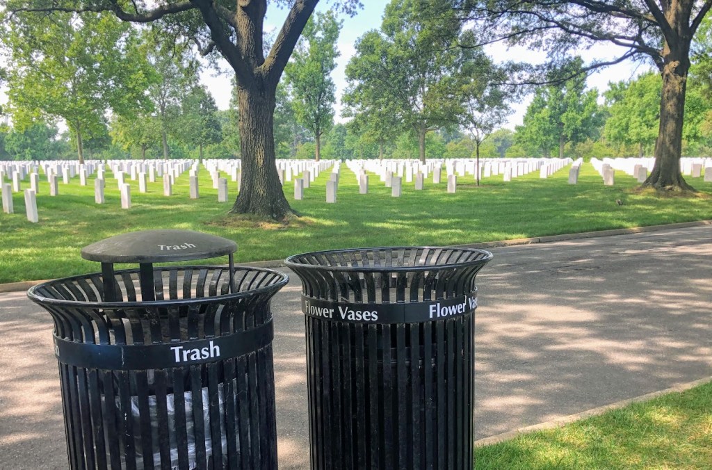Two black, metal trash cans sit at the bottom left of frame. One is marked "Trash," the other "Flower Vases." In the background, uniform rows of white grave markers fill a green lawn.