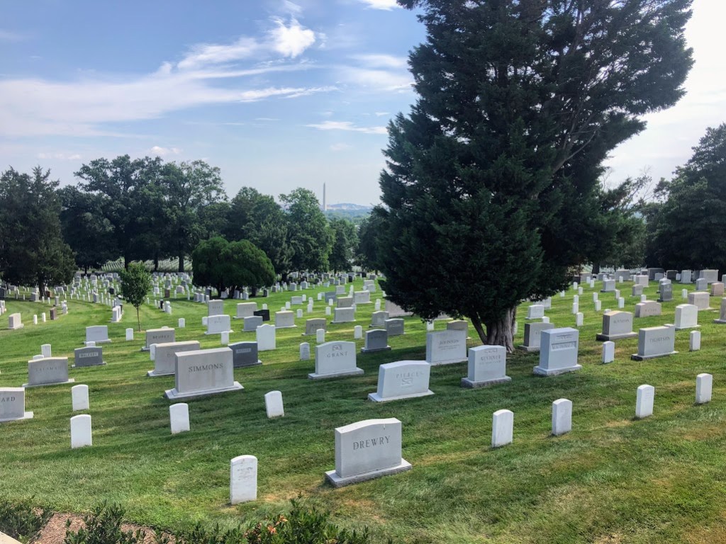 Looking down a gently sloping grassy hill covered with grave markers of many shapes and sizes. Washington Monument can be seen in the far background. 