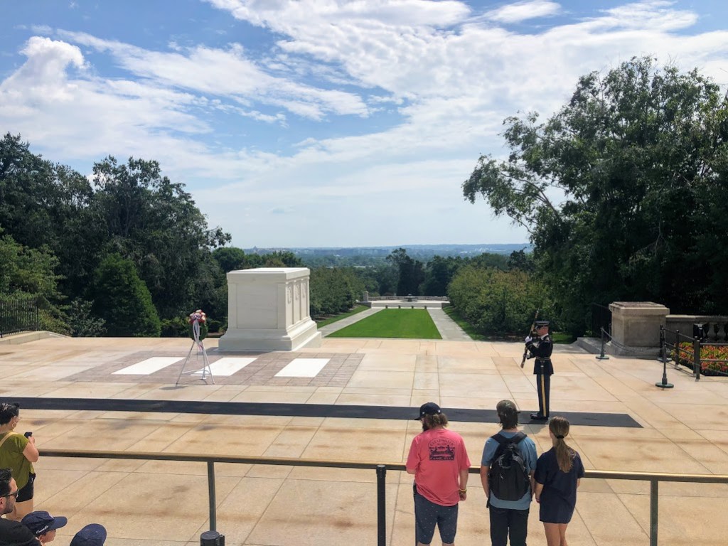 The Tomb of the Unknown Solder is at the center-left of the photo, some distance away. The Tomb Guard stands at the end of a long black track on the right side of the photo, holding his gun stiffly in front of him. Several tourists appear at the bottom of the photo, standing behind a rail and watching the solder. 