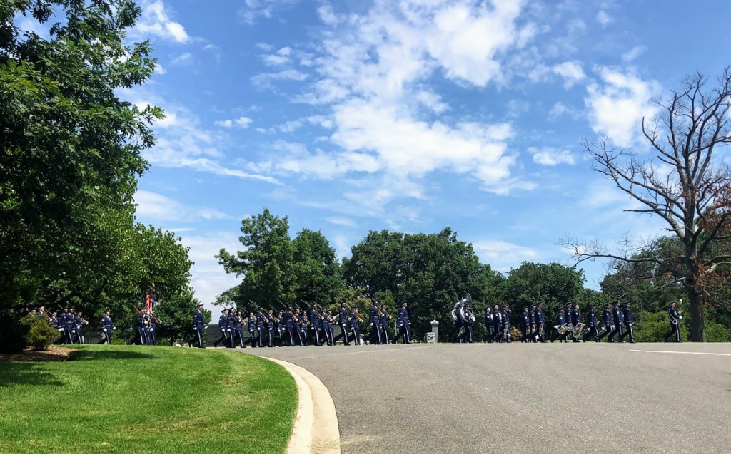 A funeral procession composed of dozens of soldiers in full military formal garb parade from the left of the frame to the right. The group of soldiers farthest to the right are playing instruments. Only the two tubas are large enough to be seen clearly. The group behind them carry guns over their shoulders. 