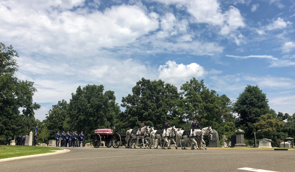 A continuation of the military funeral procession. A group of six white horses, each ridden by a soldier in uniform, pull an old-fashioned wheeled cart bearing a flag-covered casket. Two more groups of soldiers follow behind. 