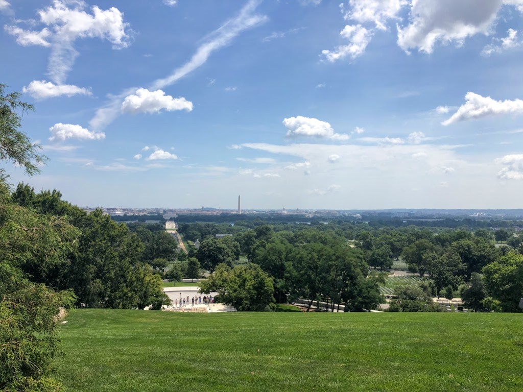 A view down a grassy slope toward a road full of tourists and farther back toward the Washington Monument in the far distance. 