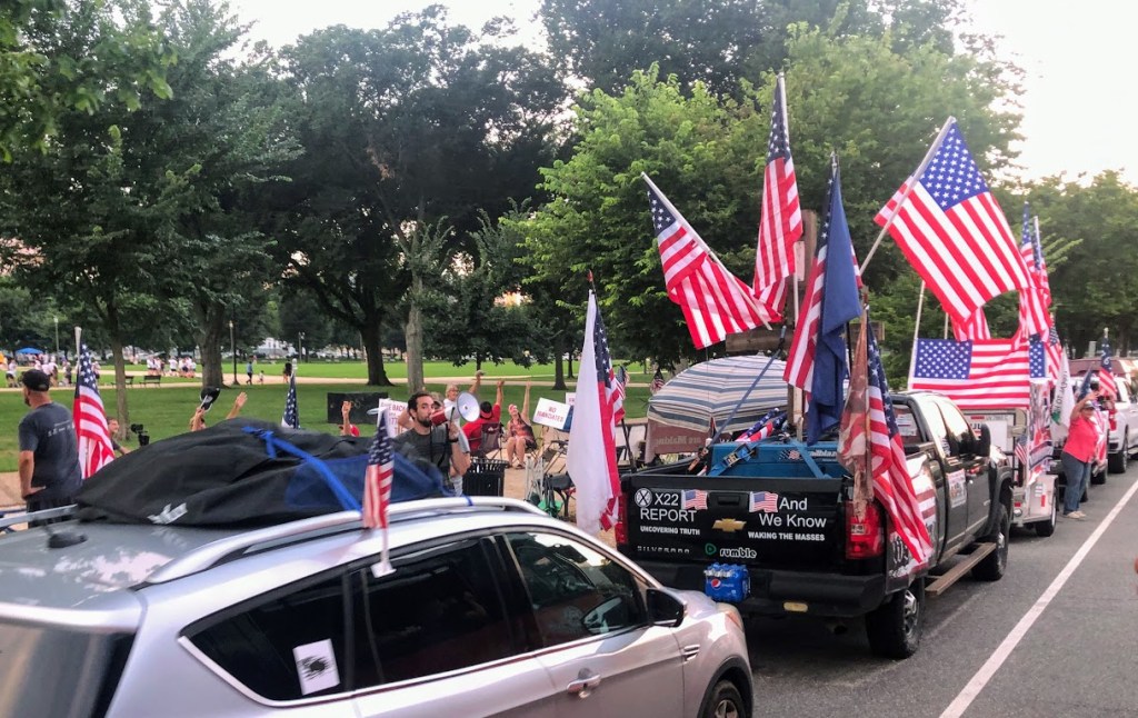 A row of vehicles is parked along the side of a grassy park. The cars and trucks are covered with more than a dozen American flags. People fill the spaces between the cars, holding signs that can't quite be read. One man speaks through a bullhorn. 