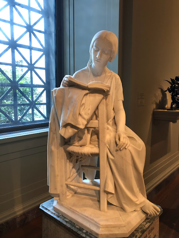 A marble sculpture of a girl sitting sideways in a chair, reading a book.
