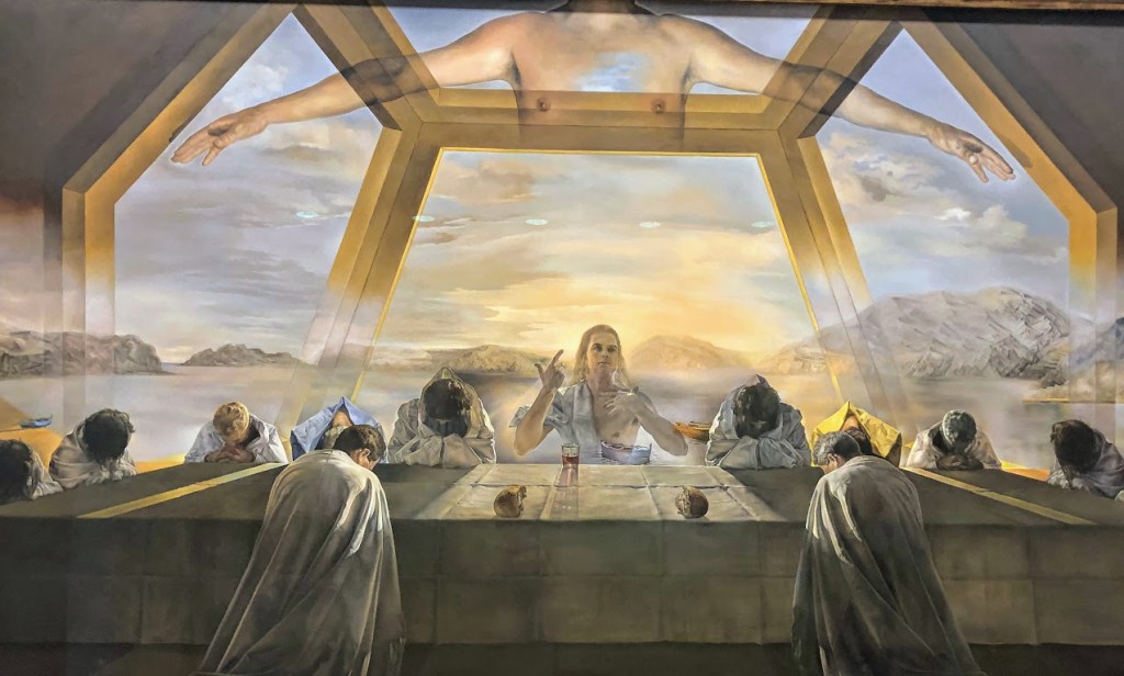 A close-up of Salvador Dali's "Last Supper." A semi-transparent Jesus sits at a table at center, framed by a large, metallic dodecahedron. His apostles pray to either side of him. The background depicts a sea-scape with cliffs and boats. A mostly transparent torso and pair of extended arms float above the whole scene. 