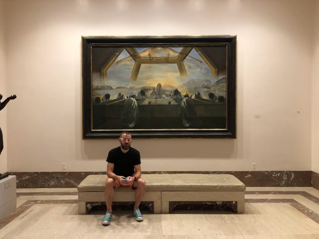 Dustin sits on a bench, bottom left, with a large, dark painting behind him. The painting is Salvador Dali's "Last Supper."
