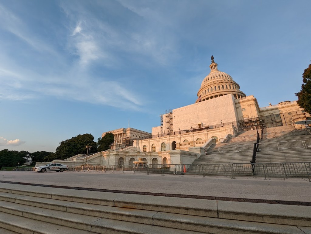 A twilight photo of the US Capitol Building with scaffolding covering most of the front, the dome rising above. A police car is parked to the far left in the foreground.