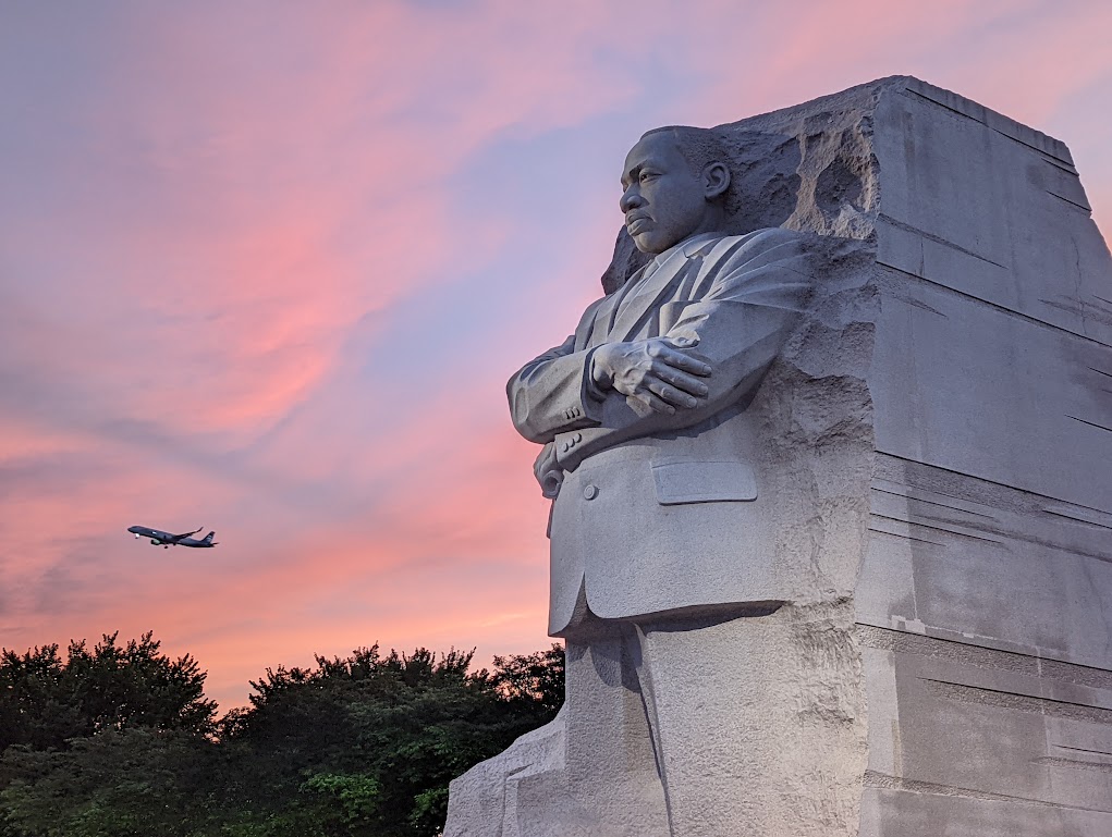 The sculpture of Martin Luther King Jr. emerging from a block of granite occupies the middle of the photo. The background shows a blue and pink sunset sky with an airplane taking off in the distance.