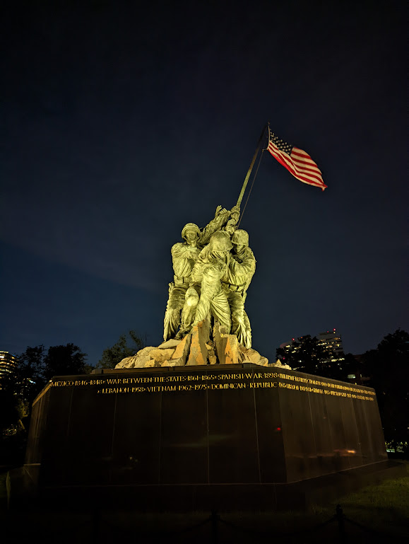 The Marine Corps Memorial fills the center of the night time photo, lit from below. This is a side-on view of the sculpture, showing the faces of the soldiers as they hoist up the flag.