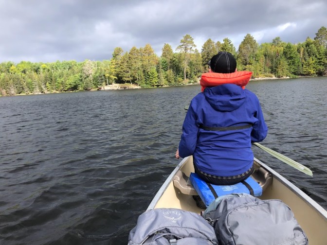 This photo is taken from the back seat of a canoe. Dustin sits in the front of the canoe with his back to the camera, wearing a blue raincoat and orange life vest. The water around him appears almost black. In the distance is a line of sunlit evergreen trees on a rocky shore. The sky above is heavily clouded.