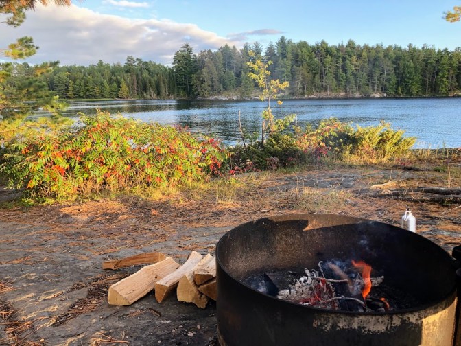In the foreground, at right, a small fire burns in a metal fire ring, though it is daytime. Cut logs are stacked to the ring's left. In the middle background, sumac bushes grow along the shore, green leaves starting to turn red. Behind them, the lake is visible. At a distance across the water you can see an opposite shore, lined with evergreen trees. The sky is blue with some gray clouds. 