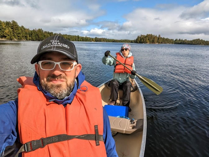A selfie taken by Dustin, who is sitting in the front of the canoe and appears large in the left of the photo. Behind him, sitting in the back of the canoe, is Laura holding a paddle at a jaunty angle and making an "ooh!" face. Both wear bright orange life vests. The lake is calm and a line of trees can be seen on the shore in the distance. 