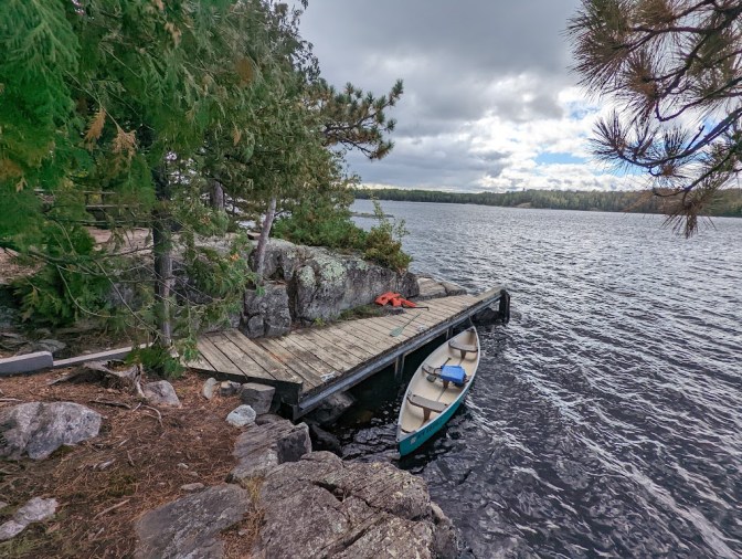 A wooden dock is built into the side of a very rocky shore. The green canoe is tied to the dock but is visibly rocking in the lake waves. The lake is dark and the sky above is cloudy.
