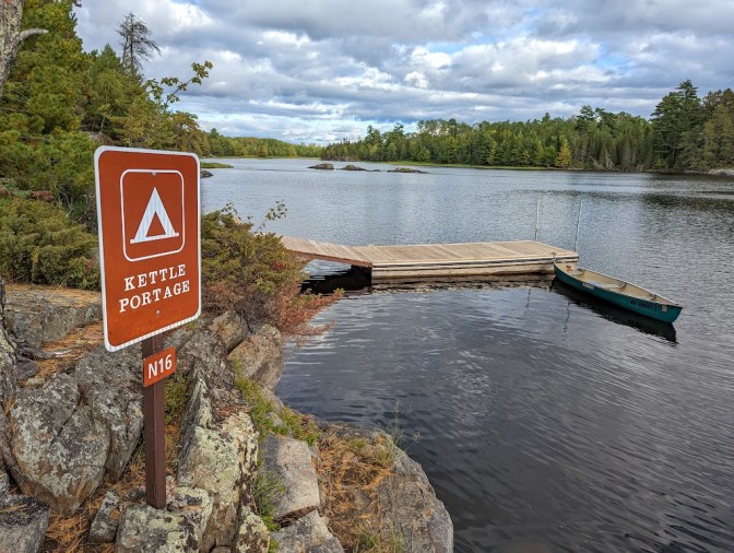 A rocky shoreline, left, holds a brown sign on a wooden post reading "KETTLE PORTAGE N16" with a tent icon, designating a campsite. A wooden dock comes out of the rocks at center, with a green canoe tied to the end. The sky above is mostly cloudy.