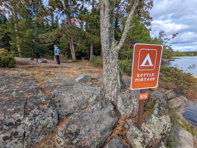 The Kettle Portage N16 campsite sign fills the right of the photo. The background looks over the campsite, lined with evergreen trees and sloping gently to the left. Laura stands in the distance in front of a bear box and beside a fire ring. 