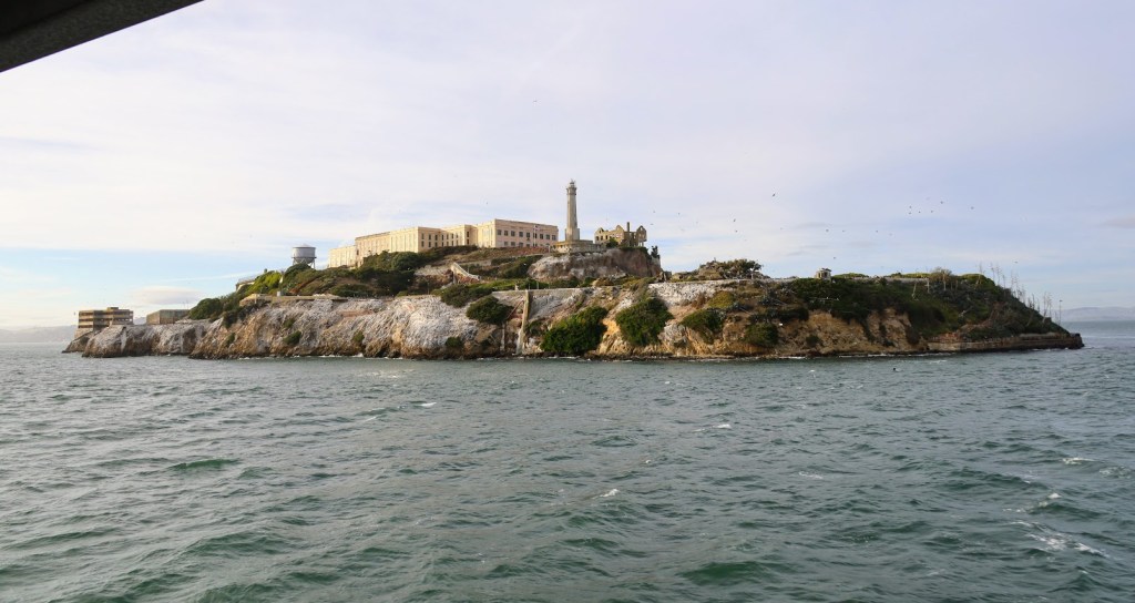 Alcatraz Island is centered in the frame. The water below is dark with a few whitecaps, the sky above cloudy. The cell block and lighthouse are visible among the rocks of the island.