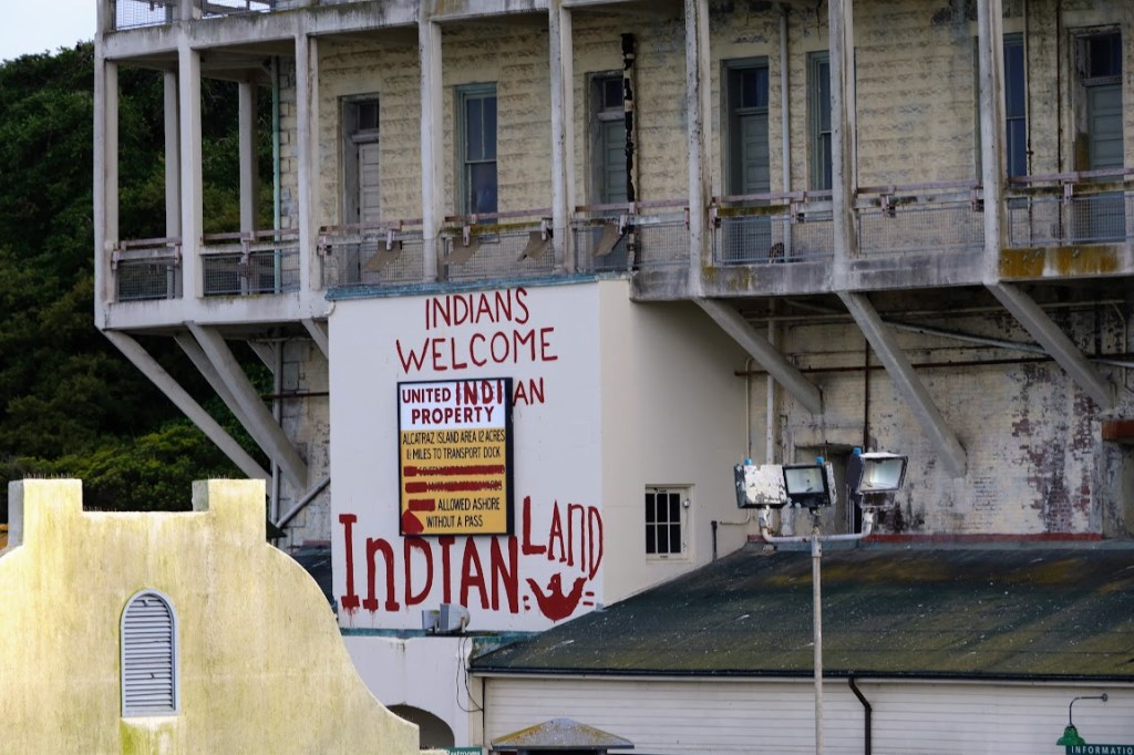 The side of a building on Alcatraz Island, its top and right side disappearing out of frame. Graffiti is painted over the top of an official government sign, reading "INDIANS WELCOME - UNITED INDIAN PROPERTY - INDIAN LAND"  A red bird with outstretched wings is also painted. 
