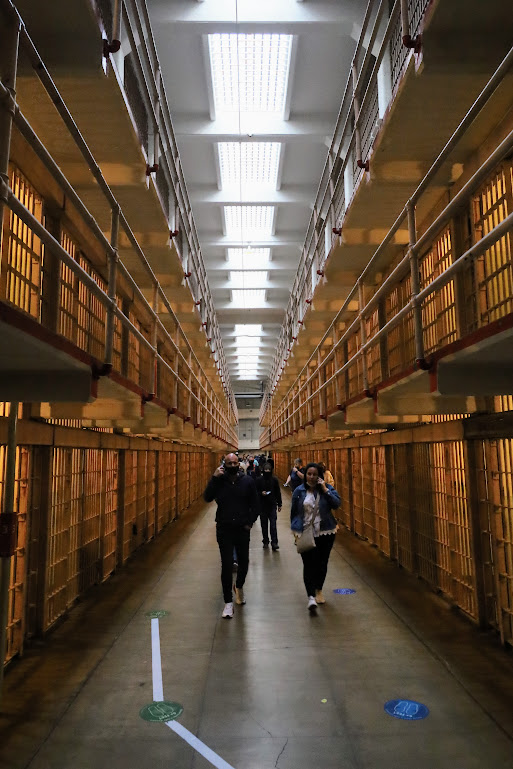 The camera looks down a three-story row of cellblocks, the aisle between them stretching an apparently long way into the distance. Lights glow orange from inside the cells while skylights above admit white daylight. The aisle is full of people listening to audiotour headsets.