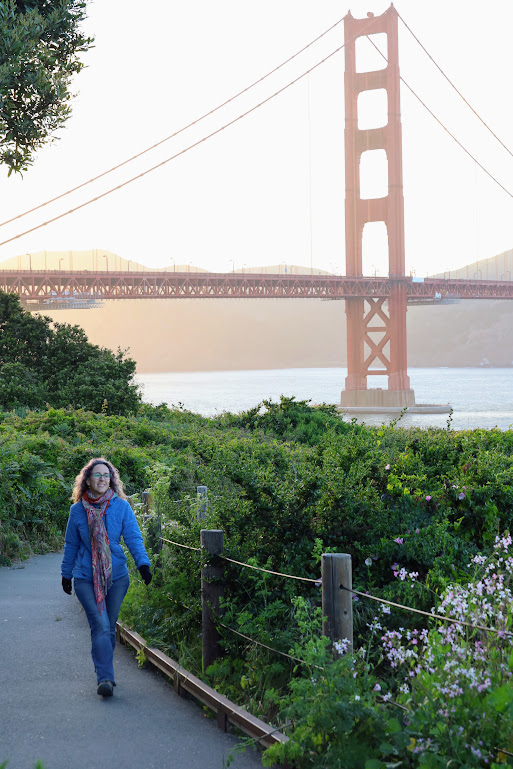 Laura walks toward the camera down a path between rows of shrubs. She strides and smiles as if she's in a catalog photoshoot. The Golden Gate Bridge rises through a slight haze in the background.