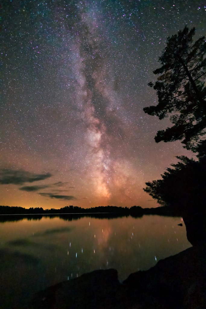 A vertical photo mostly filled with the Milky Way Galaxy stretching up from the lake. A few clouds dust the sky to the Milky Way's left. 