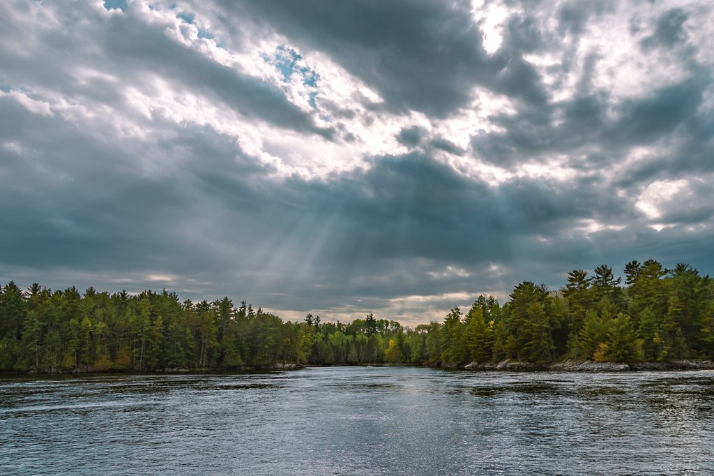 Lake at the bottom, with a tree-lined shore stretching across the middle, and a cloudy sky above. Deeply contrasted sunbeams are streaming toward the lake through breaks in the clouds above. 