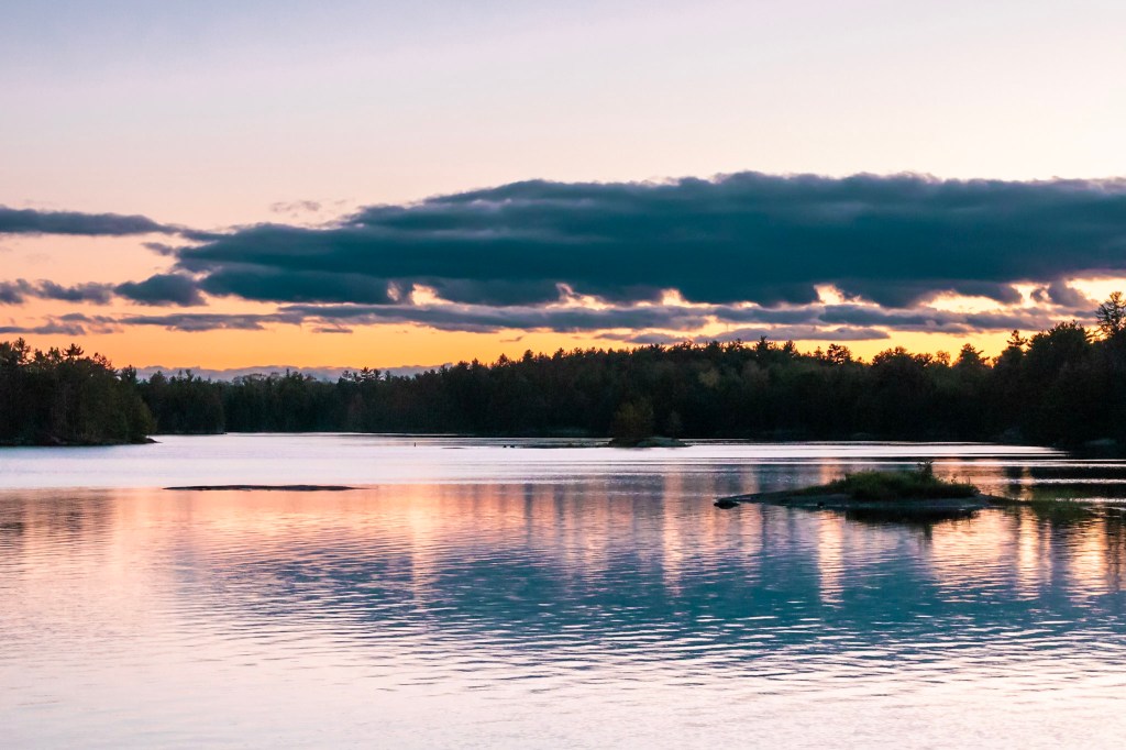 A view straight out over the lake, with a small island interrupting the water on the right. The water reflects orange light from the setting sun and a band of dark clouds.