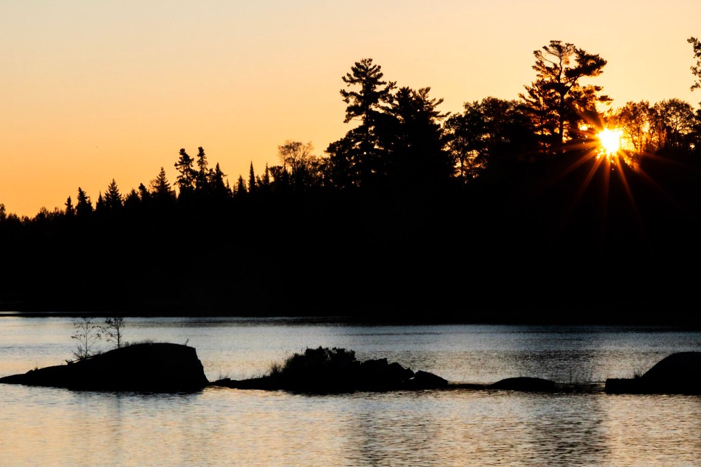 The sun appears in a starburst pattern through the silhouette of trees. The sky behind the trees is orange, and the lake below the trees is silvery. 