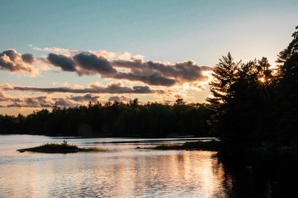 The sun creates a starburst pattern as it shines from behind the branches of evergreen trees, which appear in silhouette on the right of the frame. The lake stretches left from the trees, reflecting sunset light and bands of clouds. 