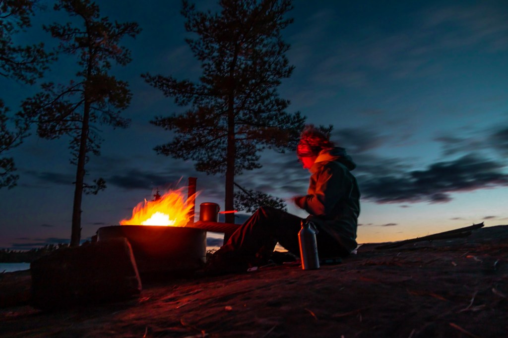 Laura sits to the right of a fire ring with a large fire burning in it. The sun has set and the sky is almost entirely dark. Laura is lit orange by the reflected firelight. 