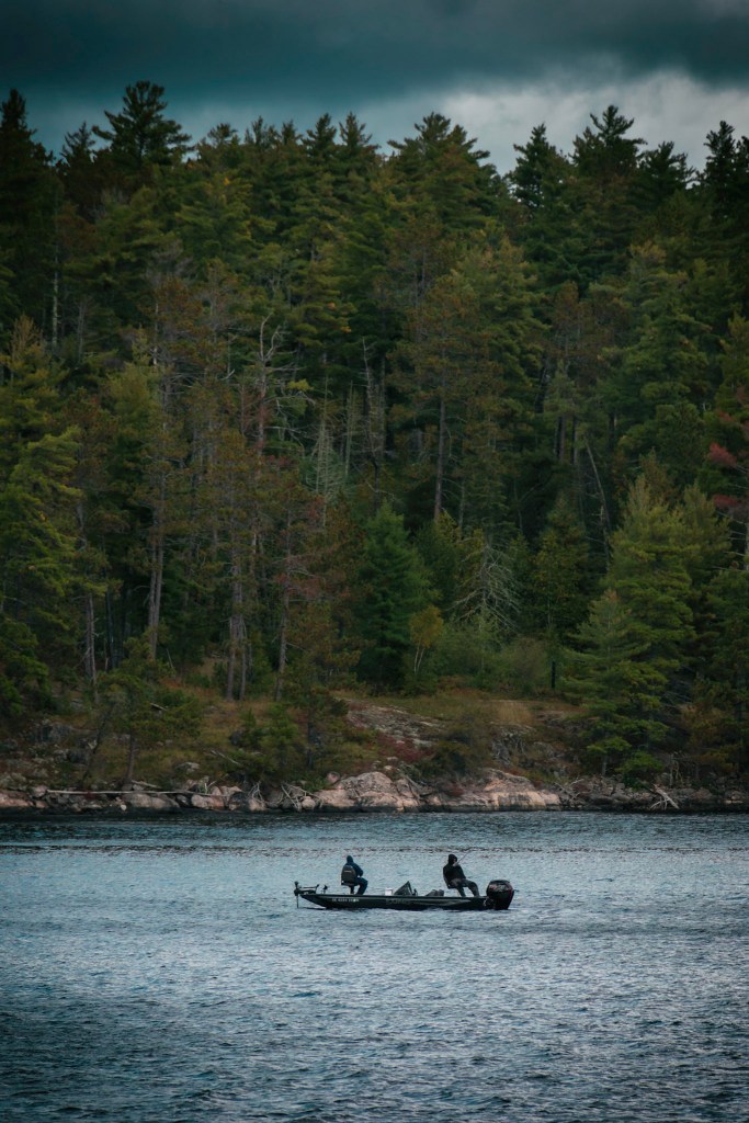 Two men sit in a boat, all appearing in silhouette against a silvery lake surface. The shore is not far behind them and is thick with evergreen trees.
