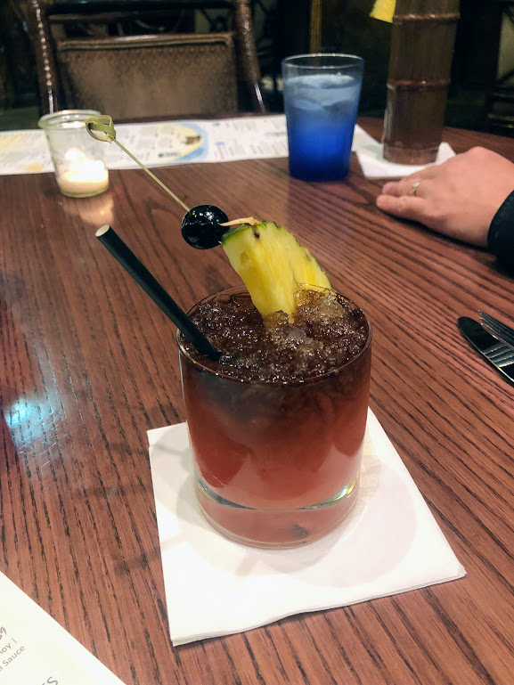 A fancy cocktail in a lowball glass that is red at the bottom and fades to near black at the top. A wedge of pineapple and dark cherry sit on a skewer at the glass's rim. 