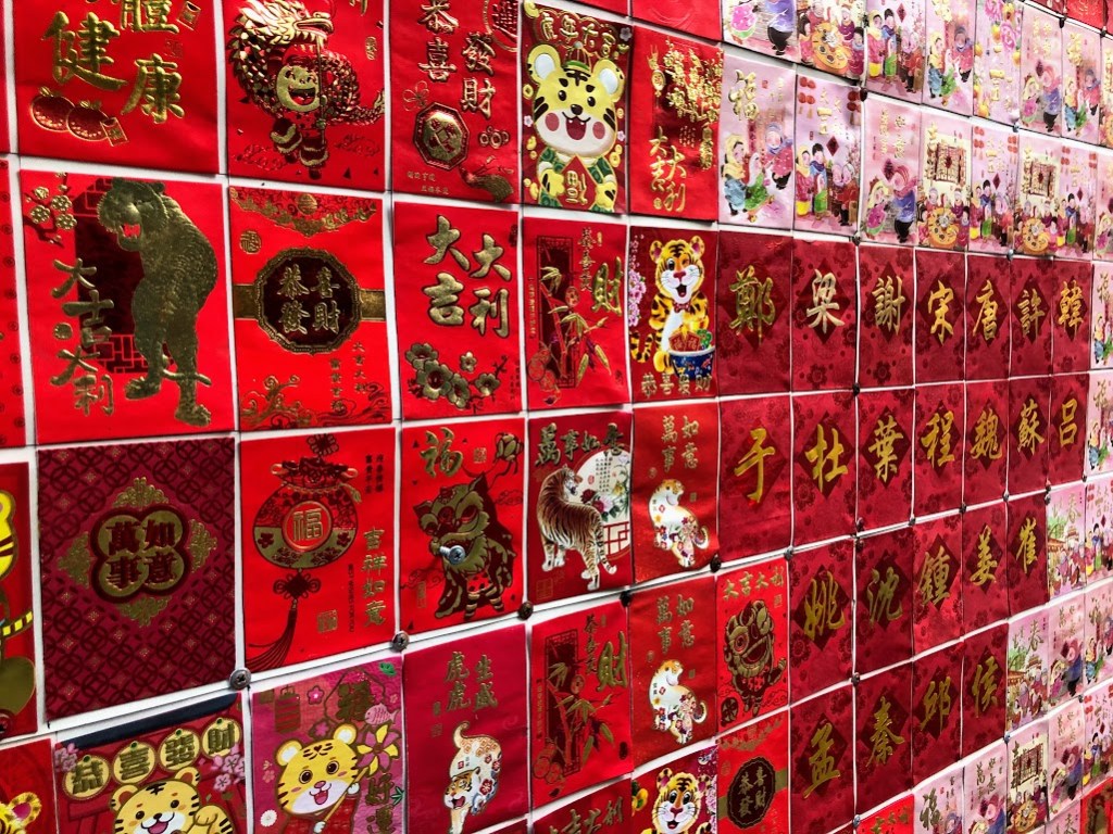 A wall is tiled in jewel-red papers decorated with gold highlights. Most depict Chinese characters. Some show cartoon tigers. 