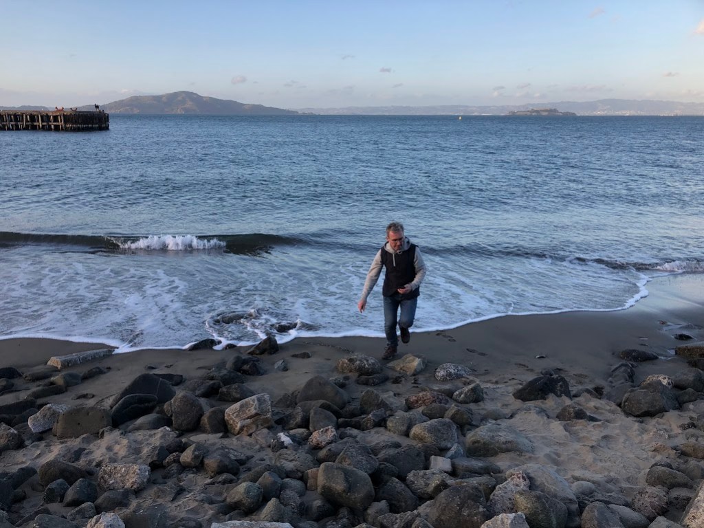 Matt retreats from an oncoming wave (a very low wave) toward rocks in the foreground of the photo. The bay stretches away behind him, with a hill rising in the far distance.