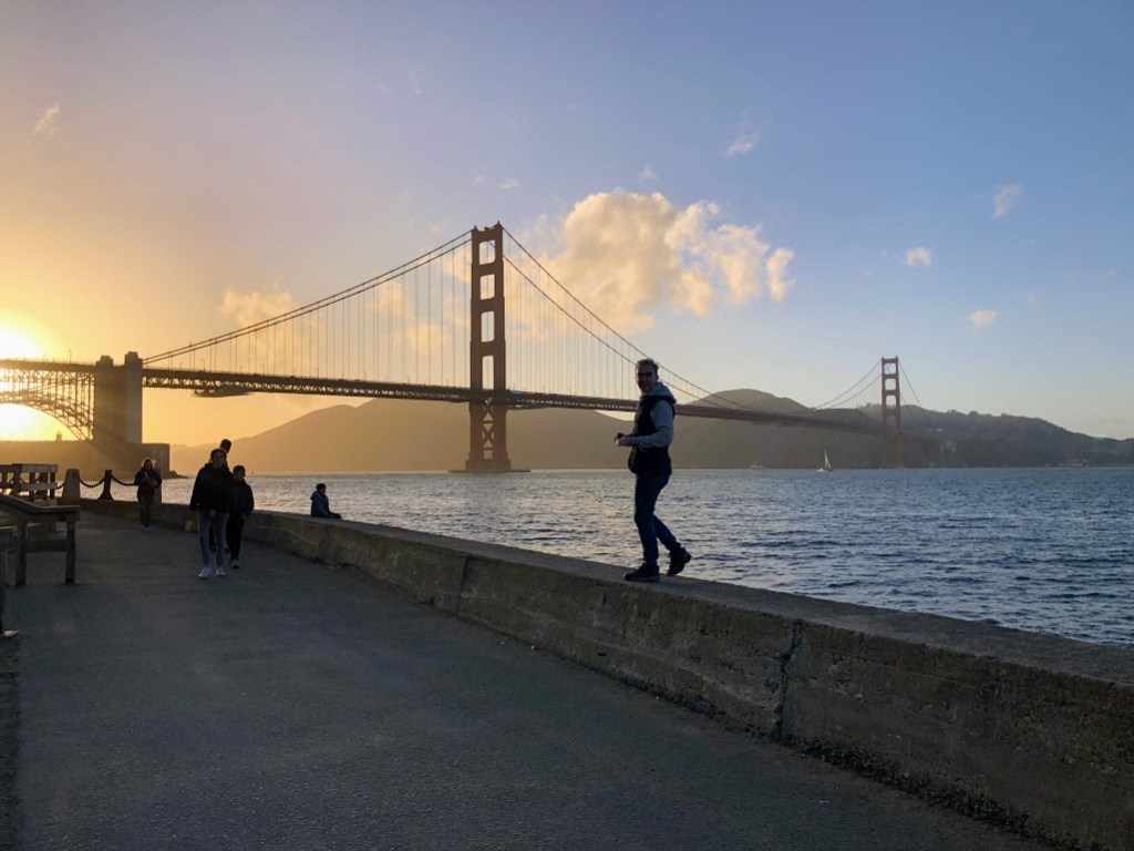 Matt walks on a low stone wall on a walkway along the San Francisco Bay. He is mostly in silhouette as the sun sets to the left side of the photo, behind the Golden Gate Bridge in the distance.