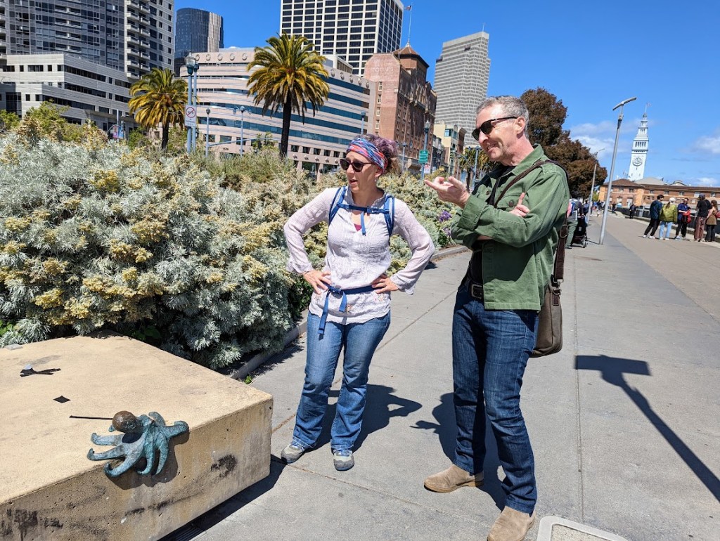 At the center of the photo, Laura stands on the left and Matt on the right. The San Francisco Embarcadero stretches out behind the, with a clocktower in the distance. They are both looking at a stone bench, on the left of the photo, which has a bronze octopus hugging its edge. 