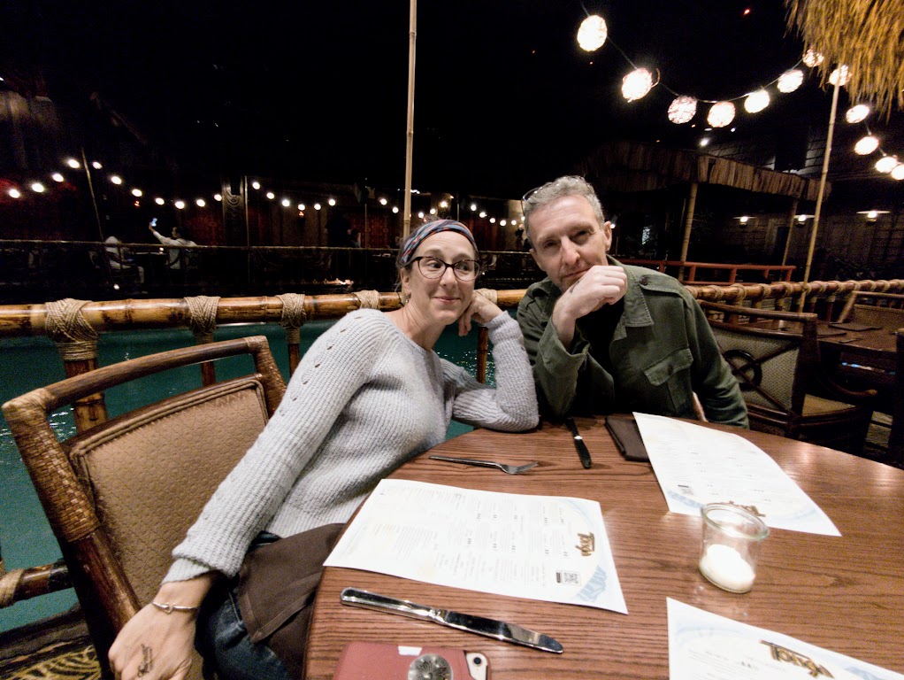 Laura and Matt sit at a restaurant table, leaning rather awkwardly together. Laura is giving Matt some suspicious side-eye. Her arm is abnormally stretched-out toward the bottom of the photo, because the perspective is a little funky. Behind their table is a bamboo railing, and behind that is a swimming pool. The room is dark with strings of globe-lights overhead.