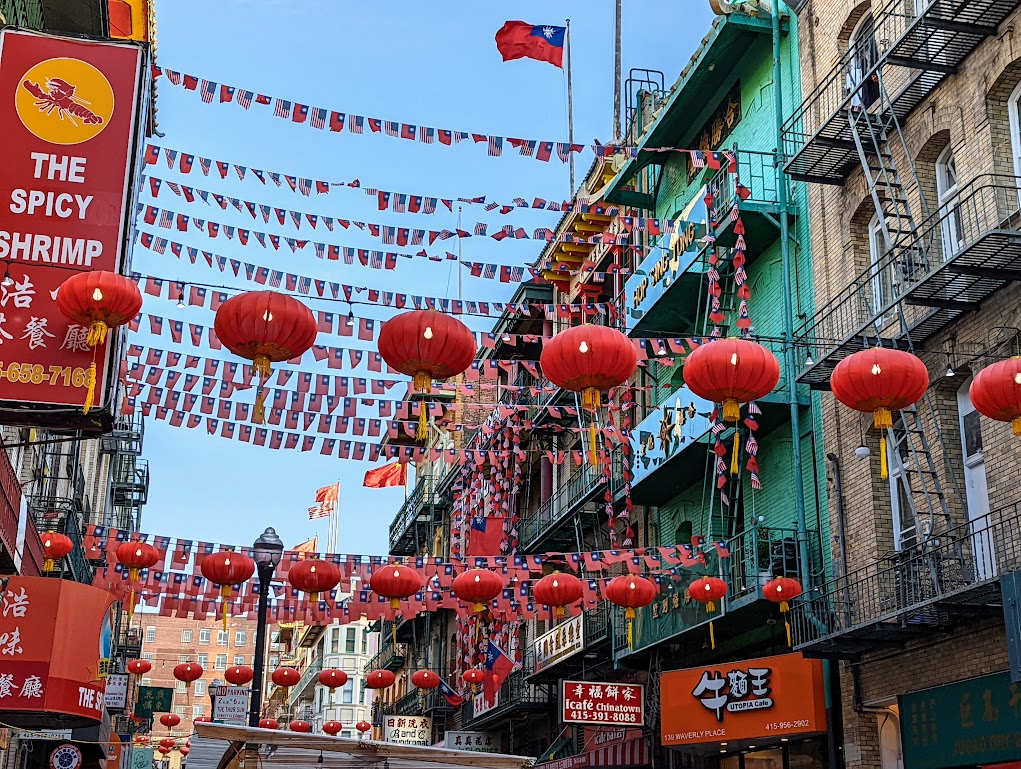 Red paper lanterns are strung across the street from building to building. On higher stories of the four-story buildings, long rows of flags are strung across. Signs on the buildings are written mainly in Chinese characters. All the buildings have balconies and fire escapes. 