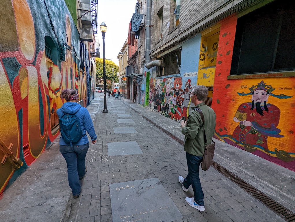 This photo looks down the length of a wide, clean alley. The walls of the alley are painted in bright oranges, reds, and other colors with graffiti showing a Chinese in traditional garb, large unreadable letters, and a parade of anime characters. Laura and Matt walk away from the camera down the alley.